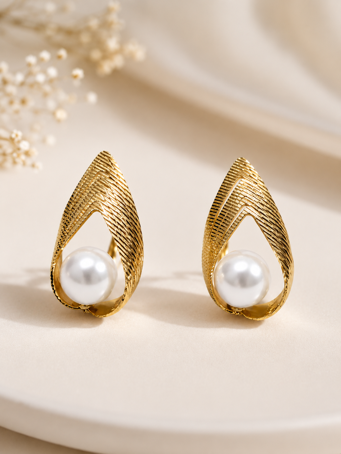 A pair of gold teardrop-shaped earrings with a textured, ribbed design and a white pearl set in the center, displayed on a light beige surface with soft, blurred dried flowers in the background.