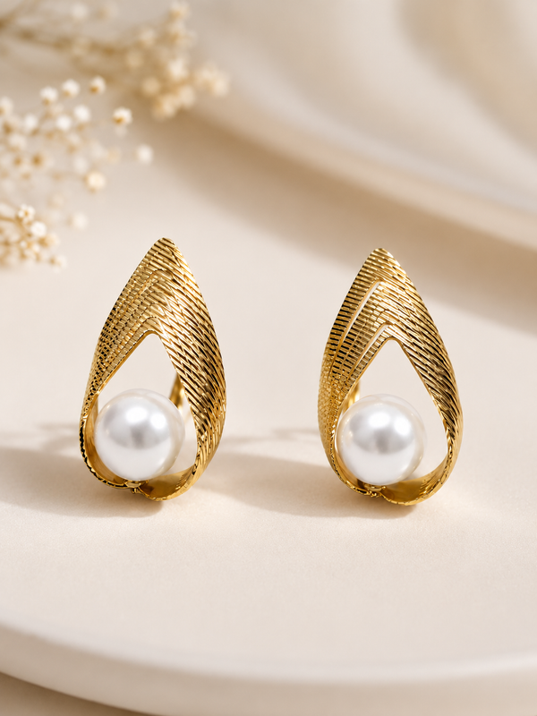 A pair of gold teardrop-shaped earrings with a textured, ribbed design and a white pearl set in the center, displayed on a light beige surface with soft, blurred dried flowers in the background.