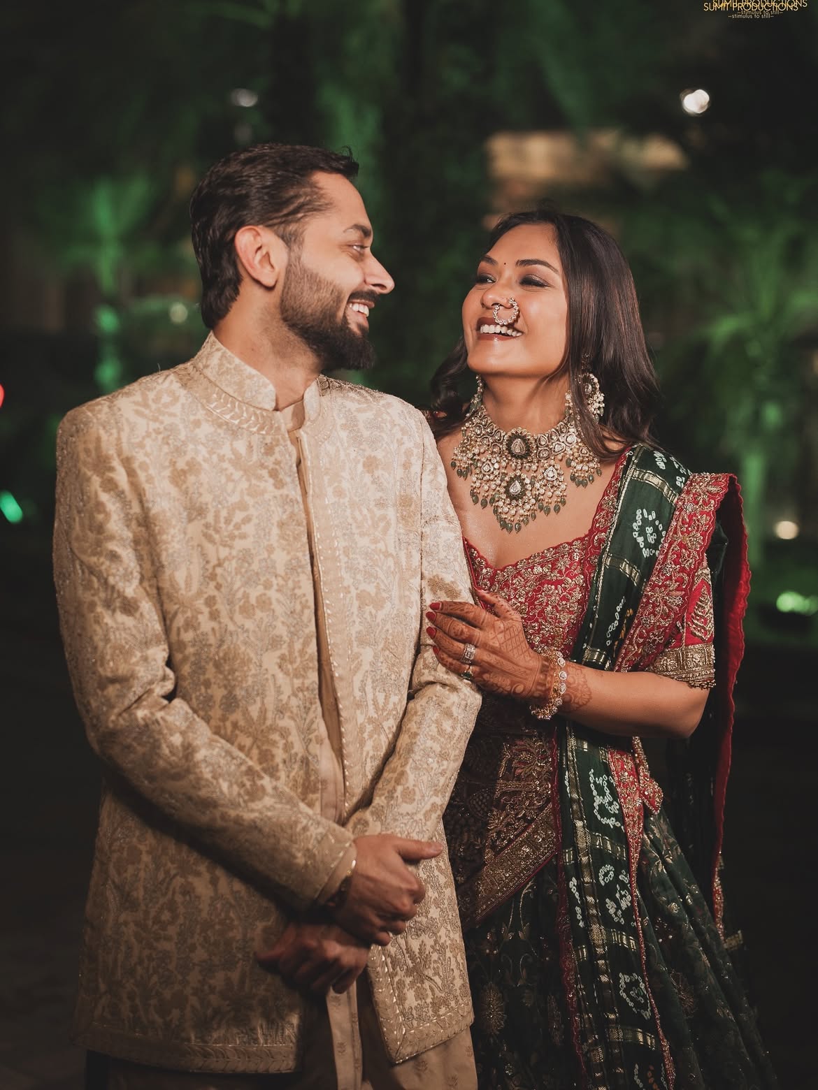 A smiling couple in traditional Indian wedding attire standing together at night. The man wears a beige patterned sherwani, and the woman wears a red and green patterned lehenga with a dark green border and ornate gold jewelry.