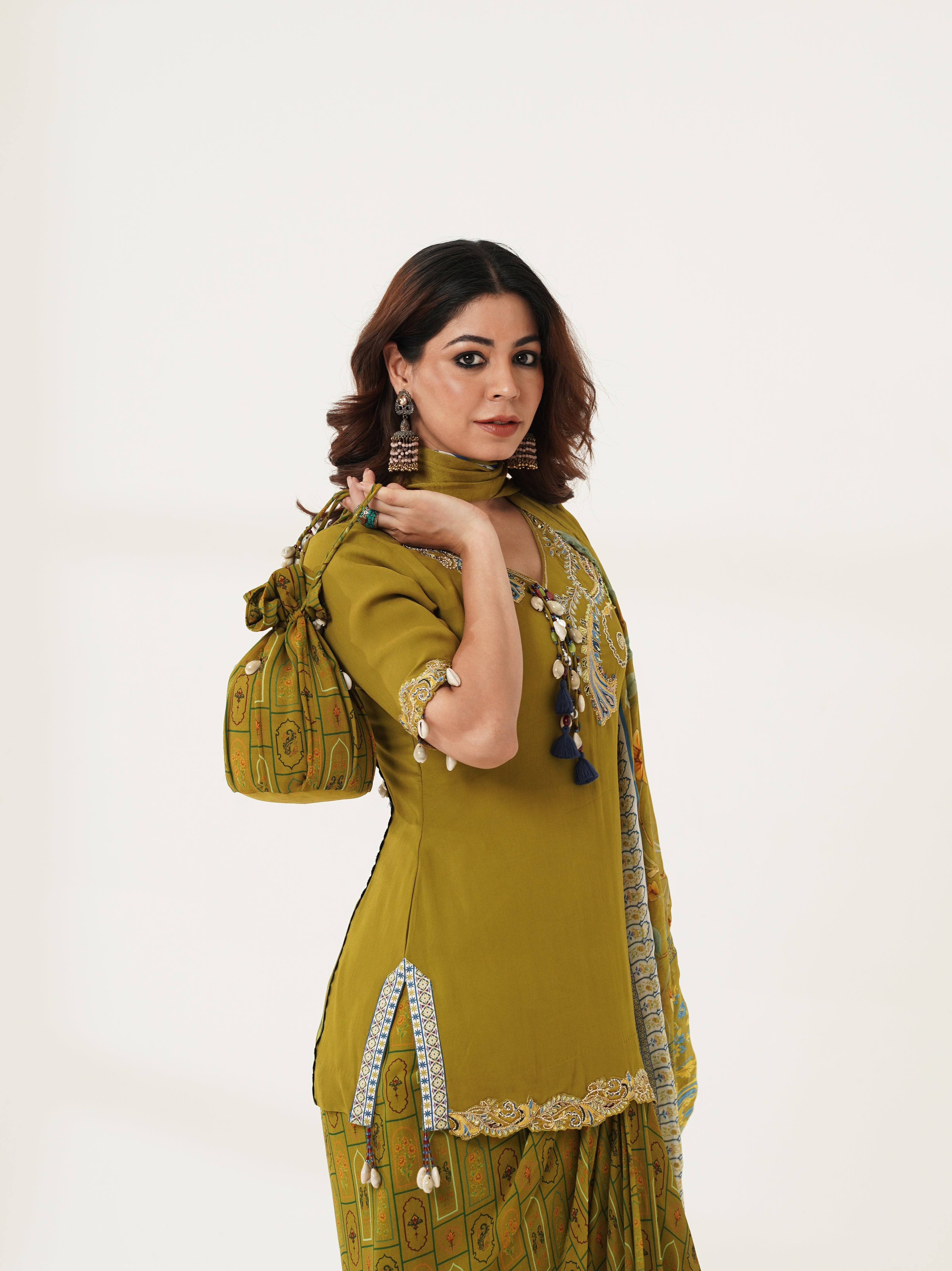 A woman modeling a traditional olive green salwar kameez set with intricate silver and gold embroidery, holding a matching patterned clutch bag against a plain white studio background.