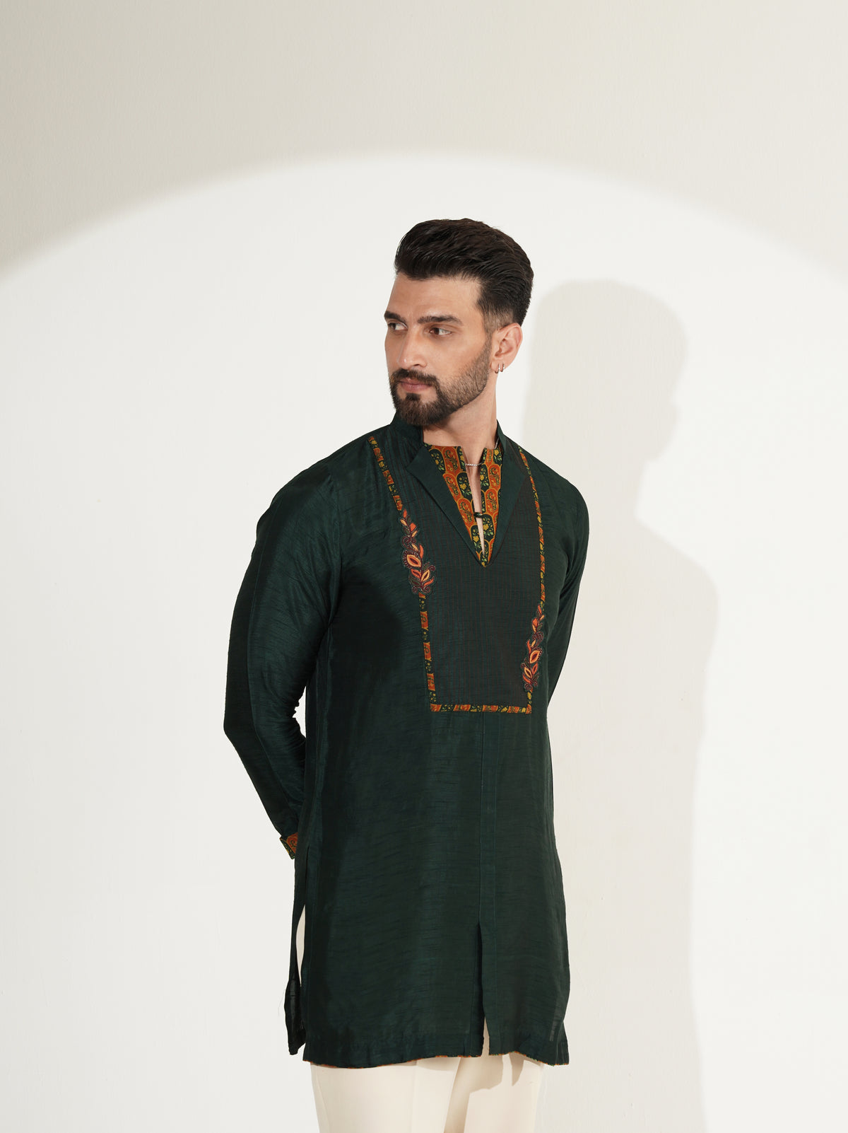 A man modeling a dark forest green long-sleeve kurta with orange and gold embroidery around the V-neck, paired with off-white trousers, set against a plain white studio background with a soft shadow.