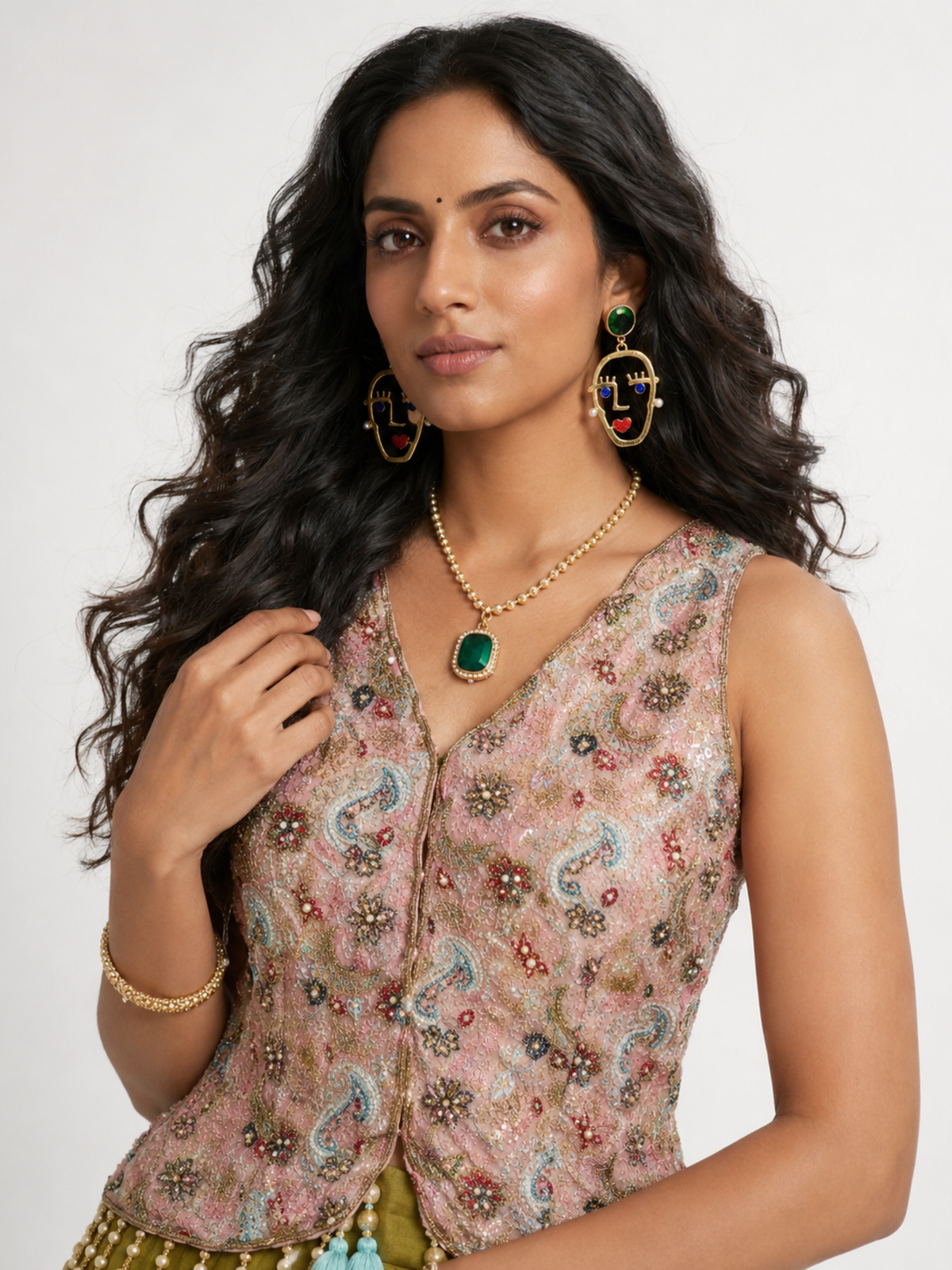 A woman modeling a sleeveless beige and pink paisley embroidered vest with gold embellishments, styled with matching gold jewelry including earrings, a necklace, and a bracelet against a plain light background.