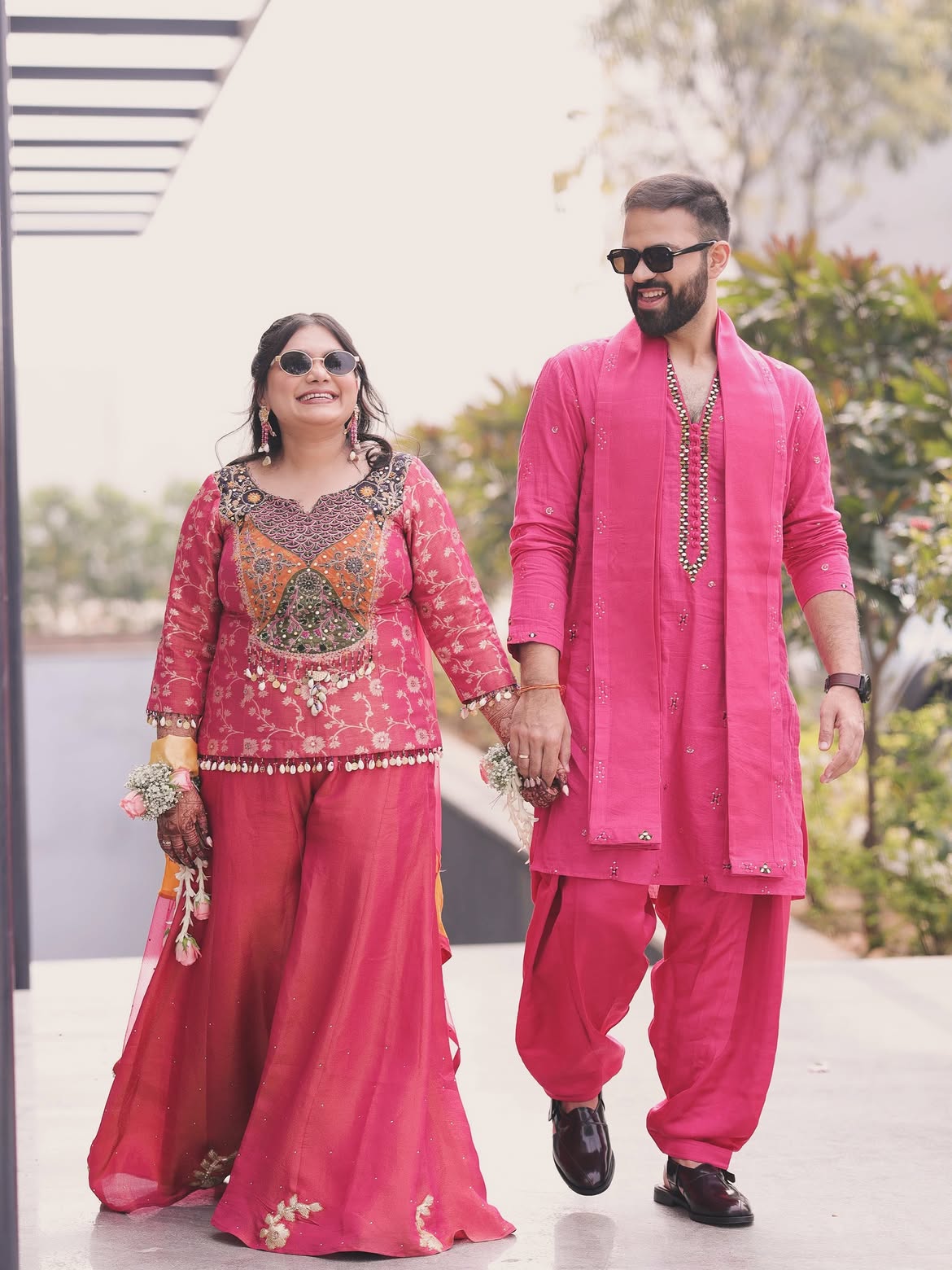 A man and a woman walking hand-in-hand outdoors. The woman is wearing a bright pink traditional outfit with intricate gold and multicolor embroidery on the bodice, paired with matching wide-leg pants.