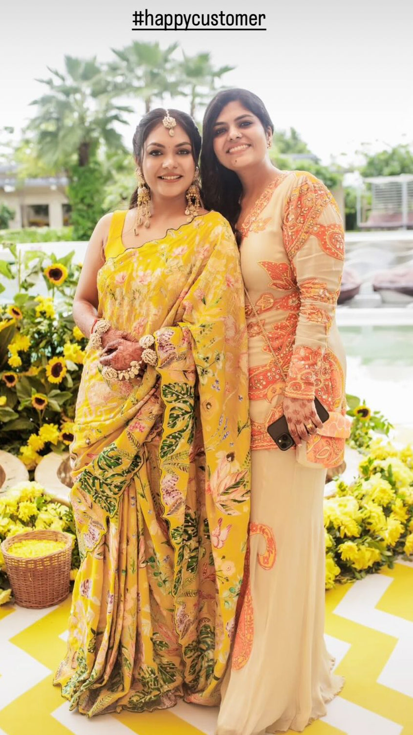 Two women posing outdoors in elegant traditional Indian attire. The woman on the left wears a vibrant yellow floral saree, and the woman on the right wears a beige kurta with orange patterns and a matching skirt.