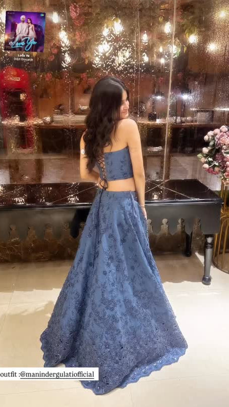 A woman wearing a two-piece blue lace formal gown with a strapless crop top and a flared skirt, photographed in an indoor event space with warm lights and a glass display case.