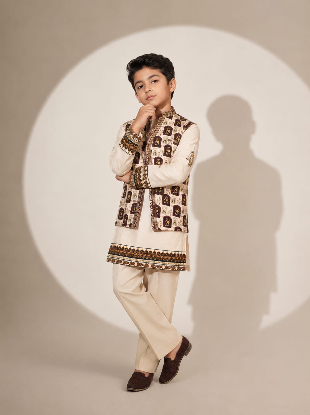 A young boy modeling a traditional Indian outfit consisting of a cream kurta with brown and gold patterned vest, cream trousers, and dark brown loafers, posed against a studio backdrop with a circular spotlight effect and his shadow.