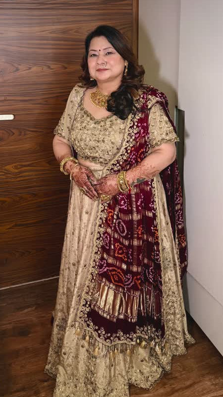 A woman wearing a traditional Indian salwar kameez outfit with a beige printed kurta and a deep maroon dupatta with gold embroidery, standing indoors in front of a wooden door.