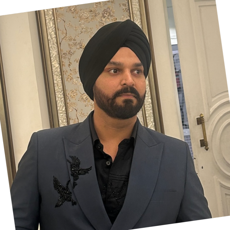 Maninder Gulati wearing a black turban and a dark gray suit jacket with a black shirt, featuring a black bird brooch on the lapel, set against an elegant indoor background.